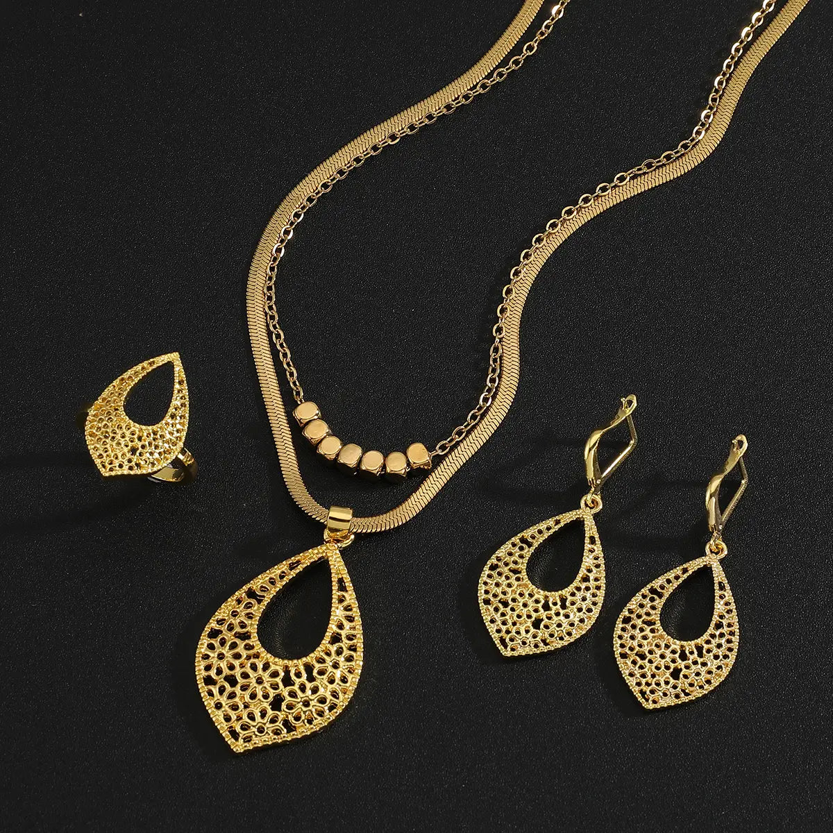 Juego de joyería de acero inoxidable color dorado para mujeres, con un collar de tono dorado con dije de forma de lágrima, aretes y anillo a juego, adornados con patrones de calado intrincado.