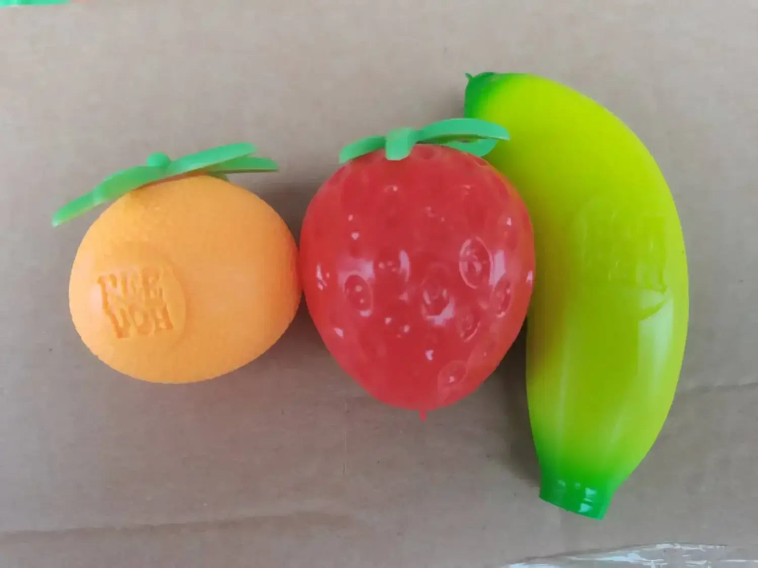 Eine Frontansicht von leuchtend orangefarbenen, roten Erdbeer- und grünen Banane-förmigen Stressbällen aus dem Erdbeere-Orange-Banane-Stressabbau-Set mit geformten Stielen.