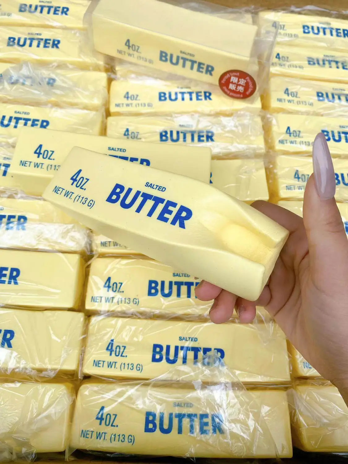 Una mano sostiene un juguete antiestrés blando con forma de barra de mantequilla salada, de color amarillo pálido, de 4 oz, que asciende lentamente, con letras azules en su envoltorio visto desde el frente.