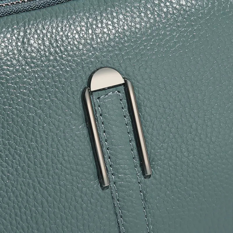 Eine Umhängetasche aus echtem Leder für Damen mit schlankem Reißverschluss-Zieher in silberfarbenem Metall, abgerundeter Oberkante und zwei vertikalen Stangen vor tealfarbenem, strukturiertem Leder.