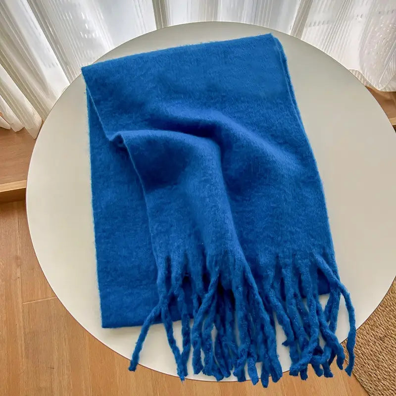 Una bufanda tejida azul real intenso con textura suave y extremos con flecos, mostrada sobre una mesa circular blanca, presentada como una elegante bufanda de cachemir de invierno para mujer.
