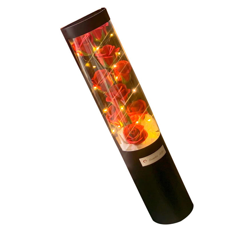 Eine zylindrische schwarze LED-Rosen-Seifenblumen-Geschenkschachtel, die rote Rosen enthält, die von warmen Feenlichtern für den Valentinstag beleuchtet werden.