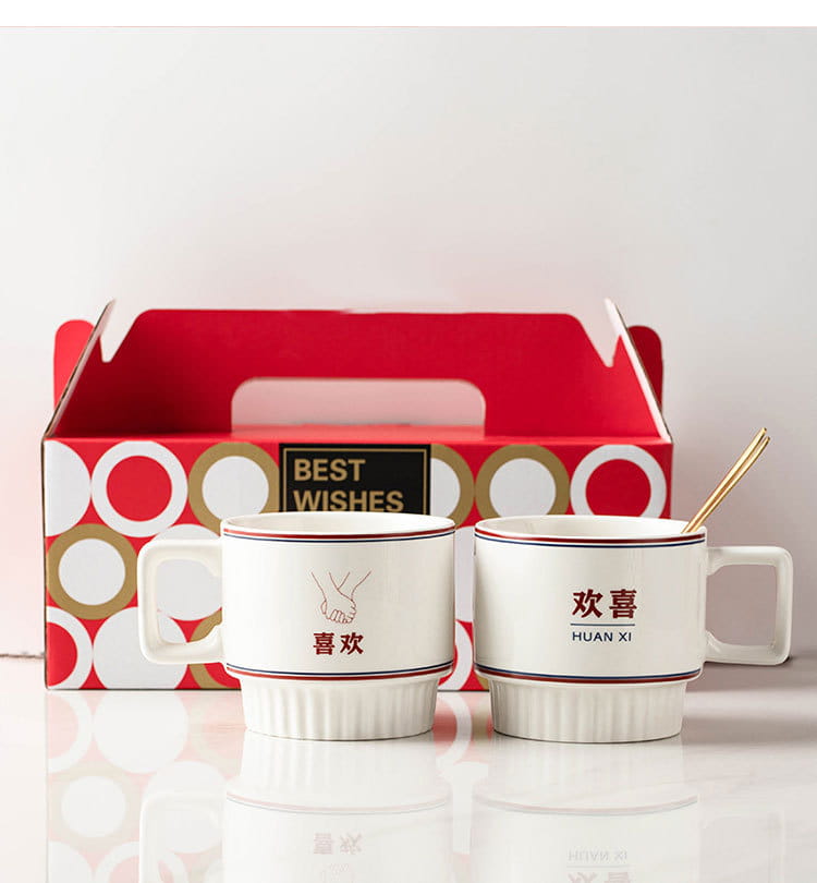 Tasses en céramique blanches avec fin liseré rouge et doré, comportant des caractères chinois « 喜 like » et « 喜 joyful », chacune accompagnée d’une cuillère assortie, dans un coffret cadeau de deux tasses de couple en céramique pour mariage chinois.