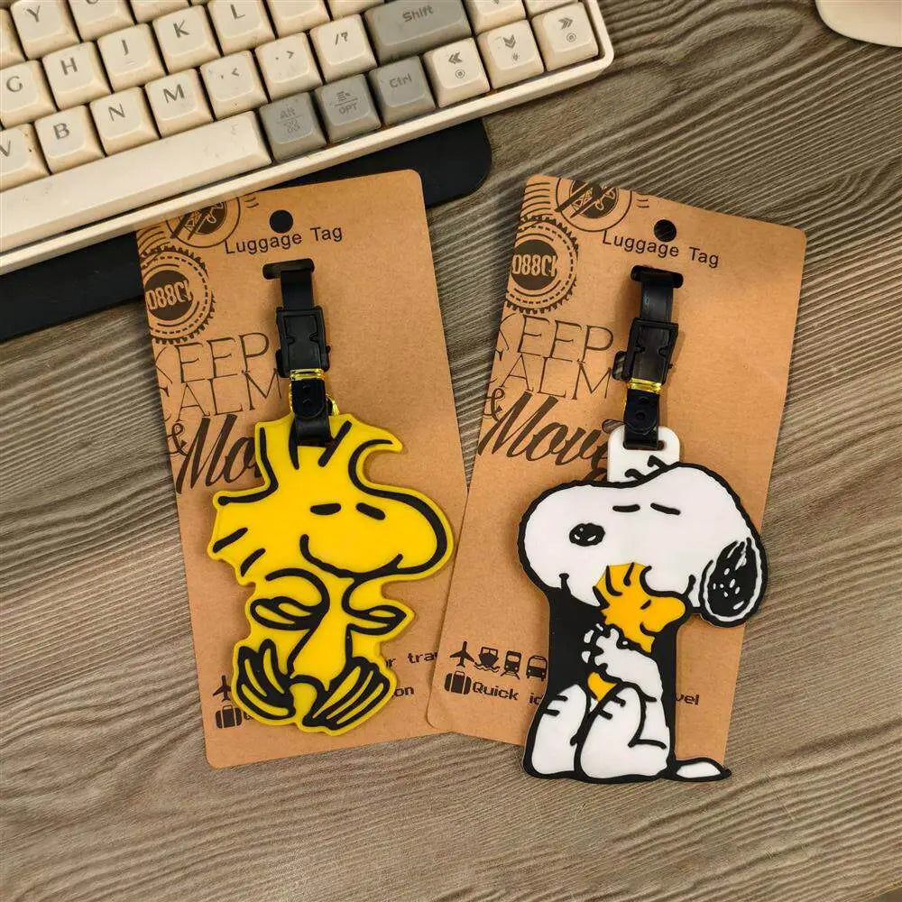 Etiqueta de equipaje de Snoopy con diseño de caricatura adorable para identificación de maleta, que presenta personajes de Snoopy y Woodstock en amarillo y blanco, con broches de plástico negros sobre un soporte de cartón.