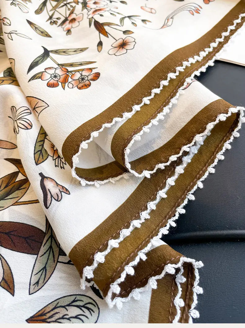 A delicate cream-colored silk floral square scarf featuring a soft peach and olive pattern with wide brown stripes and white tassel trim.