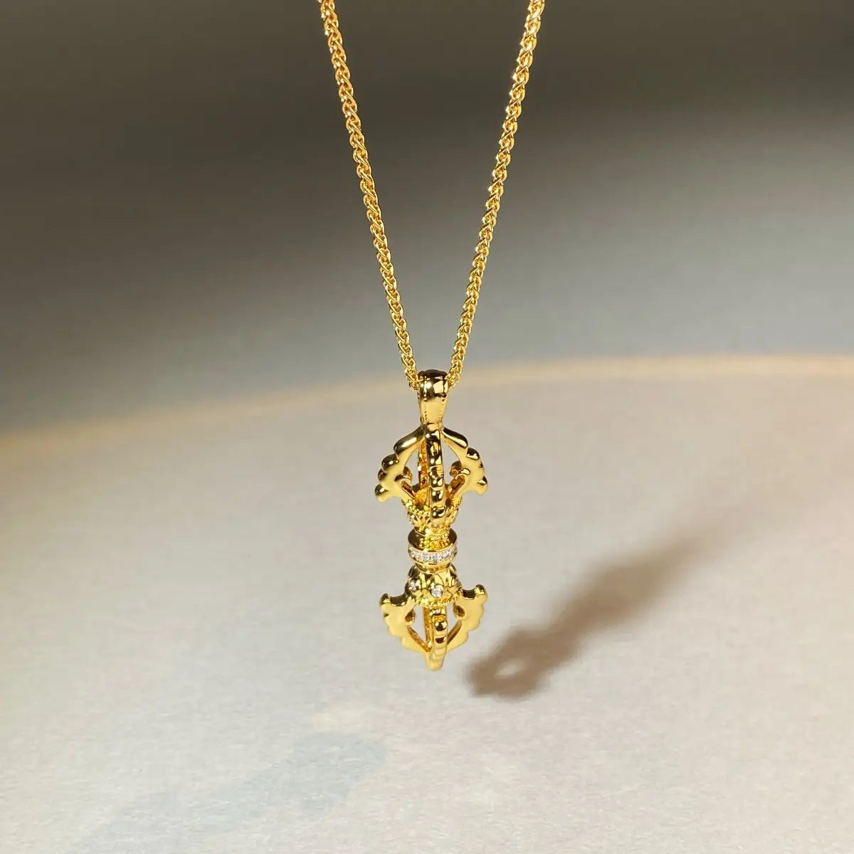 24kゴールドの金剛杵ダイヤモンド・ペンダントネックレスのデザイン。精巧な細工が施されたゴールドのチベット祈祷輪ペンダントネックレスで、その中央に小さな透明な石の列が配され、同色のゴールドチェーンに吊り下げられています。
