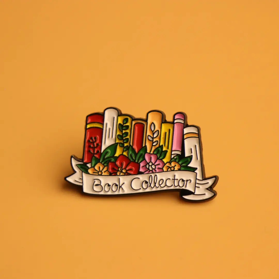 Une épingle émaillée colorée présentant des livres illustrés avec des touches florales et une bannière ornée indiquant « book collector », conçue comme broche littéraire florale.