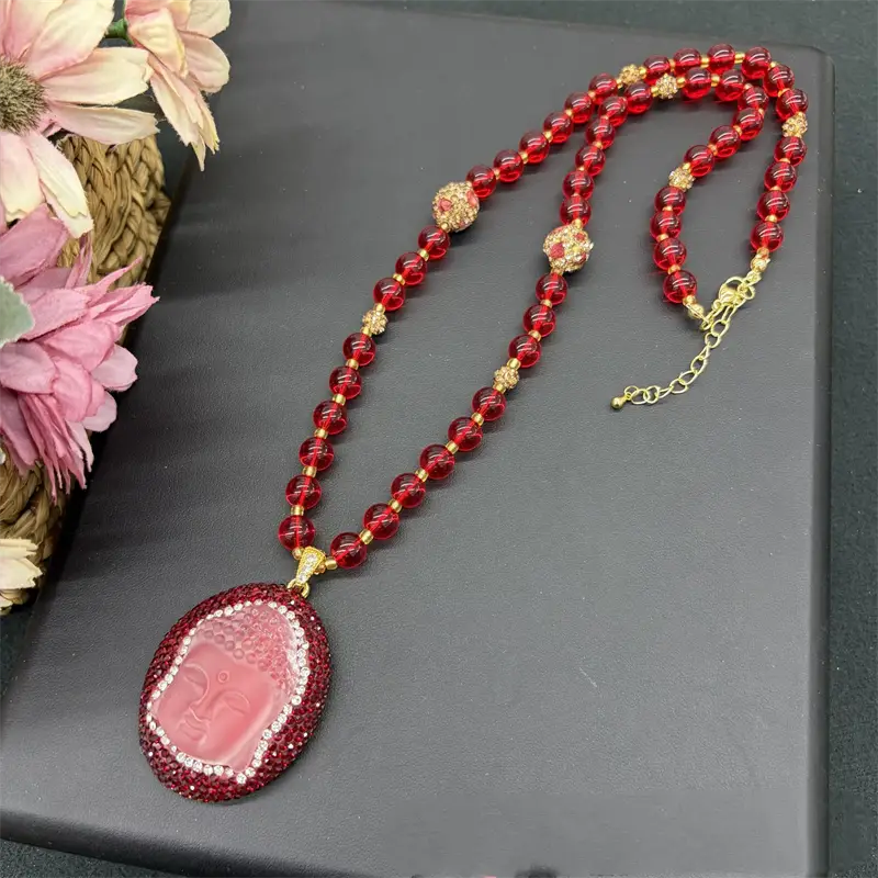 Un collier de perles rouge avec un pendentif représentant un visage de Bouddha rose, serti de strass et rehaussé d’accents ton or, adapté à divers groupes d’âge applicables et à des styles variés, y compris les accessoires à pince en forme de sésame.
