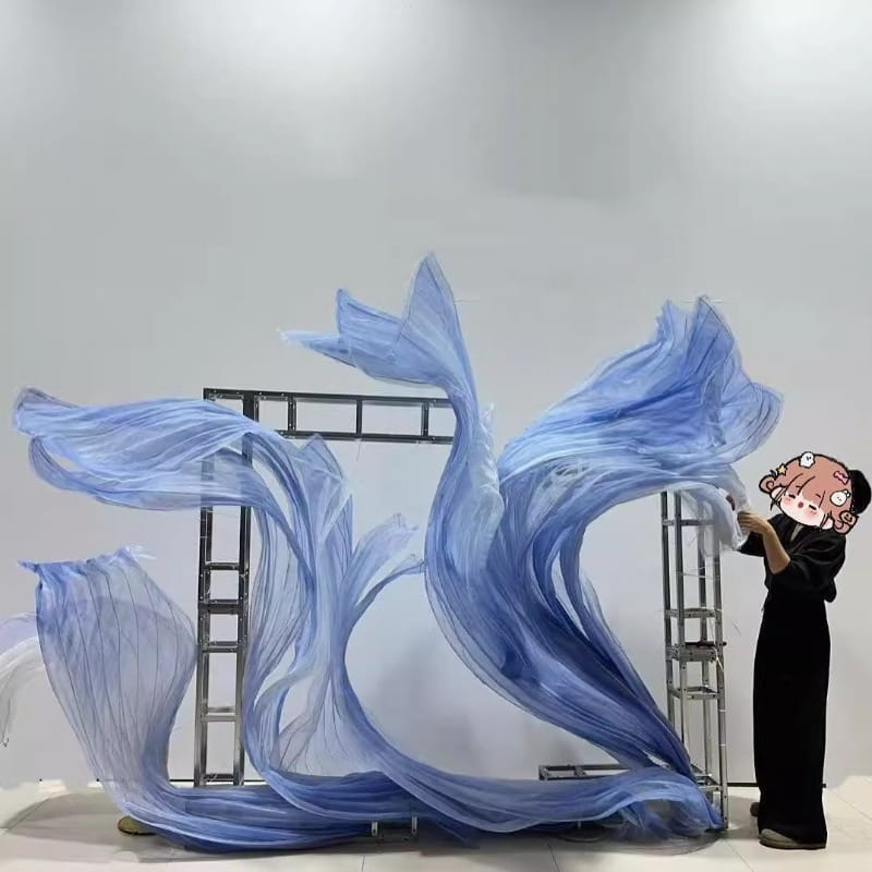 Un telón decorativo de guirnalda floral gigante de gasa que presenta un fondo decorativo con forma de flor, con una escultura de tela translúcida azul en movimiento, con pliegues dinámicos en forma de olas y un degradado de azul claro a azul profundo.