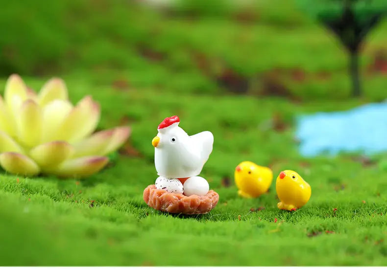 Figurine de poule en céramique blanche avec crête rouge et bec jaune, assise sur un nid orange, à côté de deux petites figurines miniatures en résine de lapin jaune, pour la décoration d’un jardin féerique.