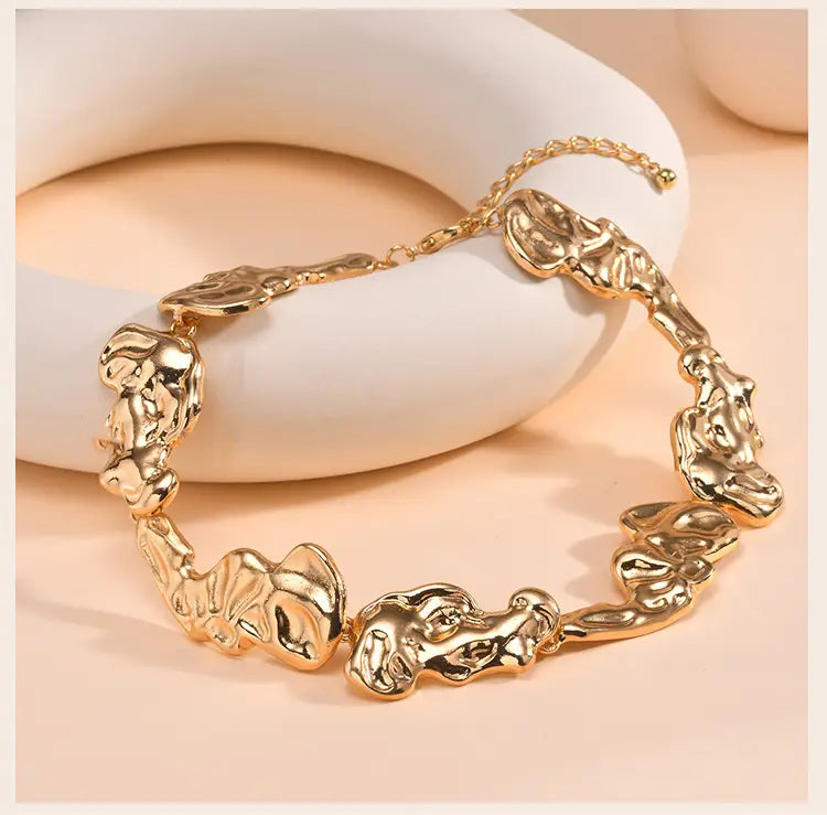 Bracelet ton or présentant des breloques sculpturales abstraites à l’aspect de visage avec une finition métallique texturée, complétant les bijoux de ras-du-cou minimaliste en lave dorée.