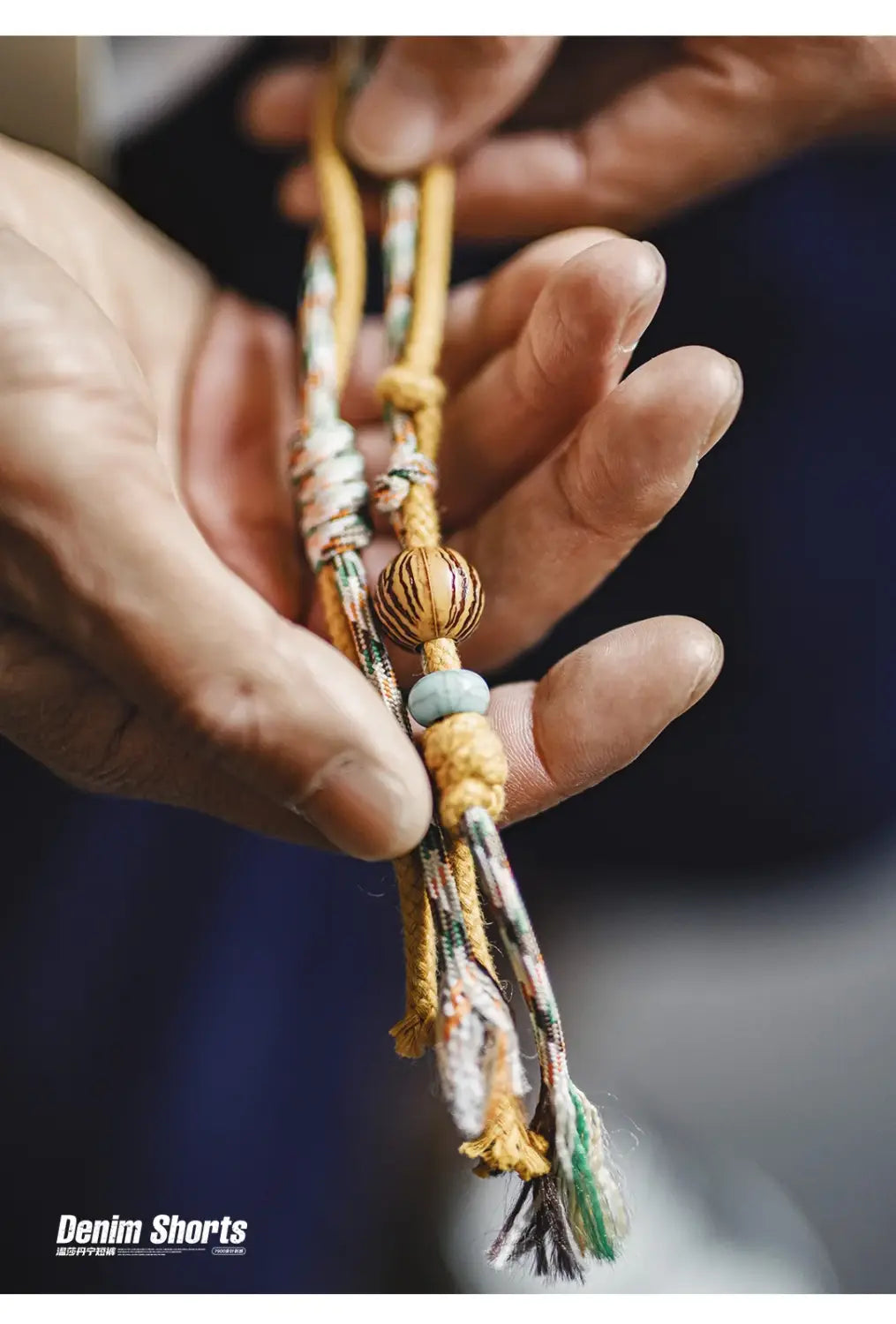 Handgefertigte Perlenkette mit einem zentralen, gestreiften Holzperlen-Element, einem hellblauen jadeartigen Spacer sowie mehrfarbigen, gewebten Bandenden in Kombination mit Vintage bestickten Jeansshorts für Herren.