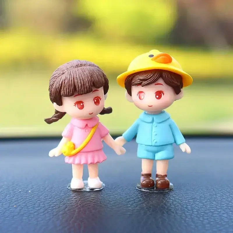 ピンクの女の子とブルーの男の子のプラスチック製フィギュアが手をつないでおり、男の子が黄色いアヒルの帽子をかぶっている様子をあしらった、かわいいカップルのカー・ダッシュボード用レジンオーナメント。