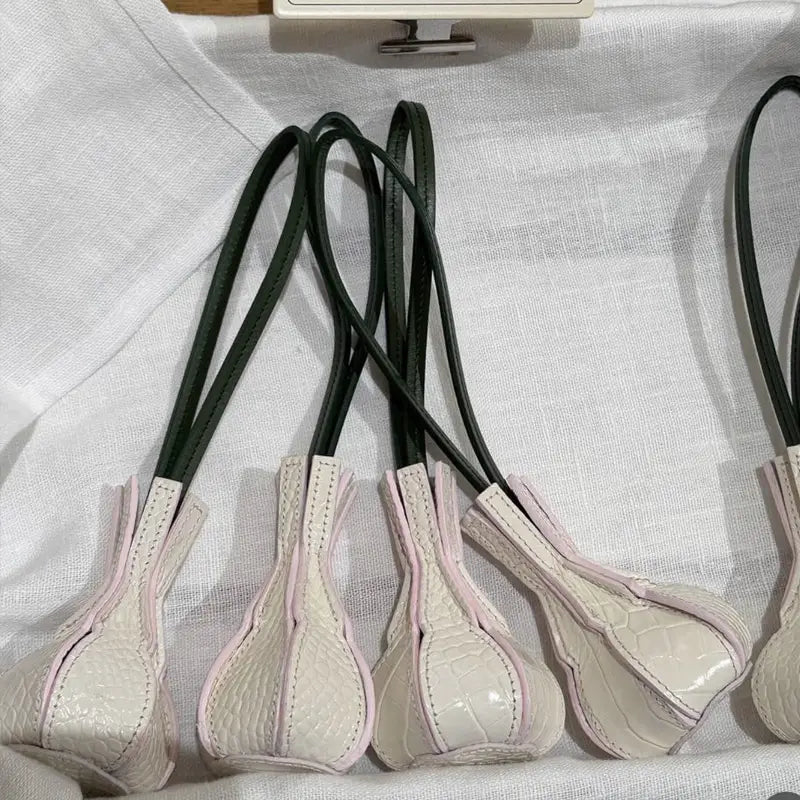 Des lanières de sac à main en cuir végétal gaufré façon crocodile, couleur crème, avec des poignées noires et une couture rose, conçues comme charms et ornements de sac à main de créateur.
