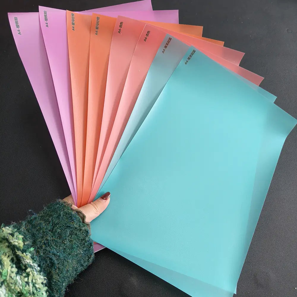 Una mano sostiene un manojo de carpetas de plástico translúcidas tamaño A4 en tonos pastel de rosa, naranja, durazno y turquesa, utilizadas para organizar documentos relacionados con el costo de envío y los accesorios de la máquina de laminación al vacío de película para teléfonos móviles.
