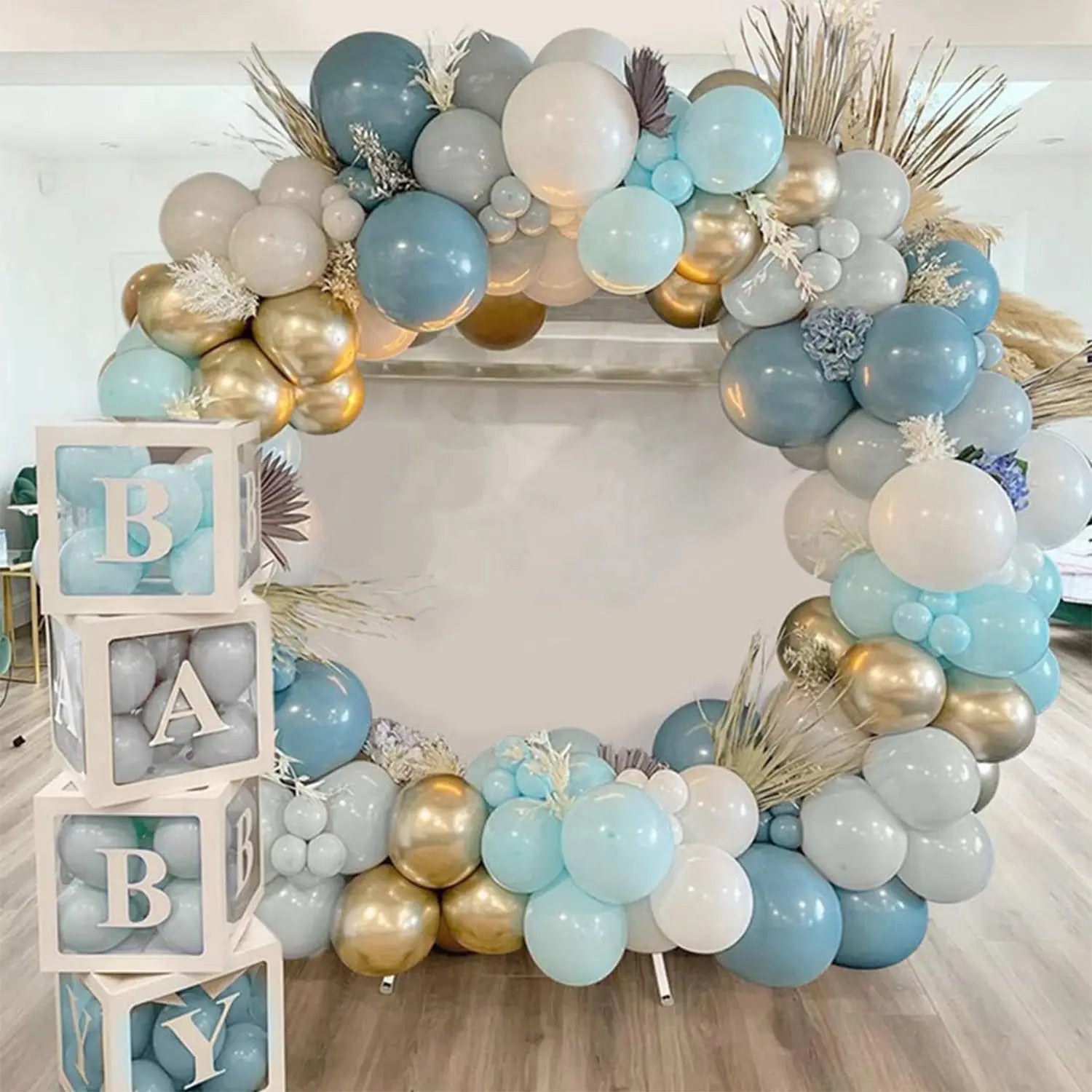 Ein dekorativer Ballonbogen mit hellblauen, weißen und goldenen Ballons sowie Akzenten aus getrocknetem Gras ergänzt die Baby-Dusch-Serviettenballonboxen, 4-teiliges Set transparenter Buchstabenblöcke.