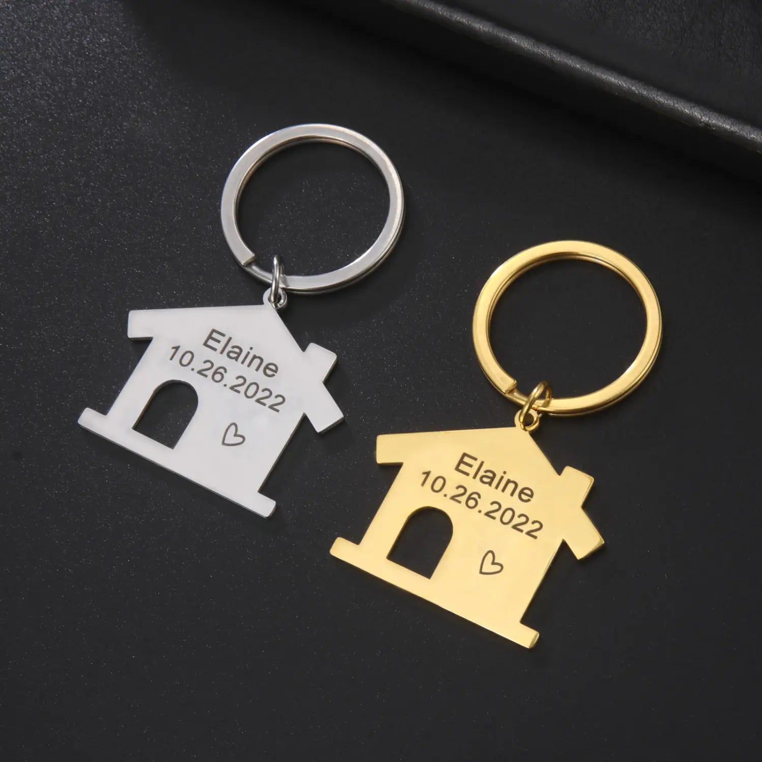 Two house-shaped personalized keychains in silver and gold metal engraved with the name Elaine and date 10.26.2022 with a small heart beneath each, ideal as a custom couple gift.