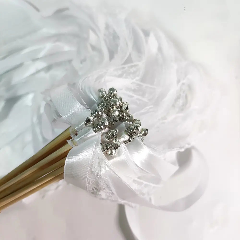 Une baguette de ruban de mariage comportant un délicat ruban en satin blanc noué autour d’un bouquet de bâtonnets, agrémenté d’un élégant ensemble de perles métalliques argentées, rehaussé de dentelle et de cloches