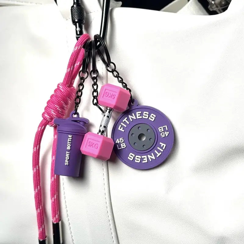 Un porte-clés de fitness violet et rose comprenant des breloques d’haltères en silicone, un pendentif de bouteille shaker et une breloque de disque de poids pour la décoration d’un sac de sport.