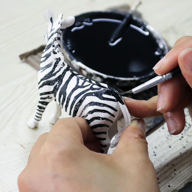 A hand-painted white zebra figurine with bold black stripes being detailed with a brush as soft animal figurine decompression toys finished goods.
