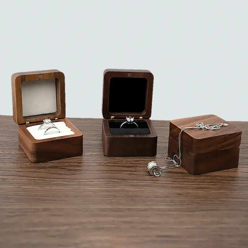 Trois présentoirs carrés en bois pour bague d’engagement et proposition de mariage, dont un avec un intérieur blanc et l’autre avec un intérieur noir, contenant tous deux des bagues.