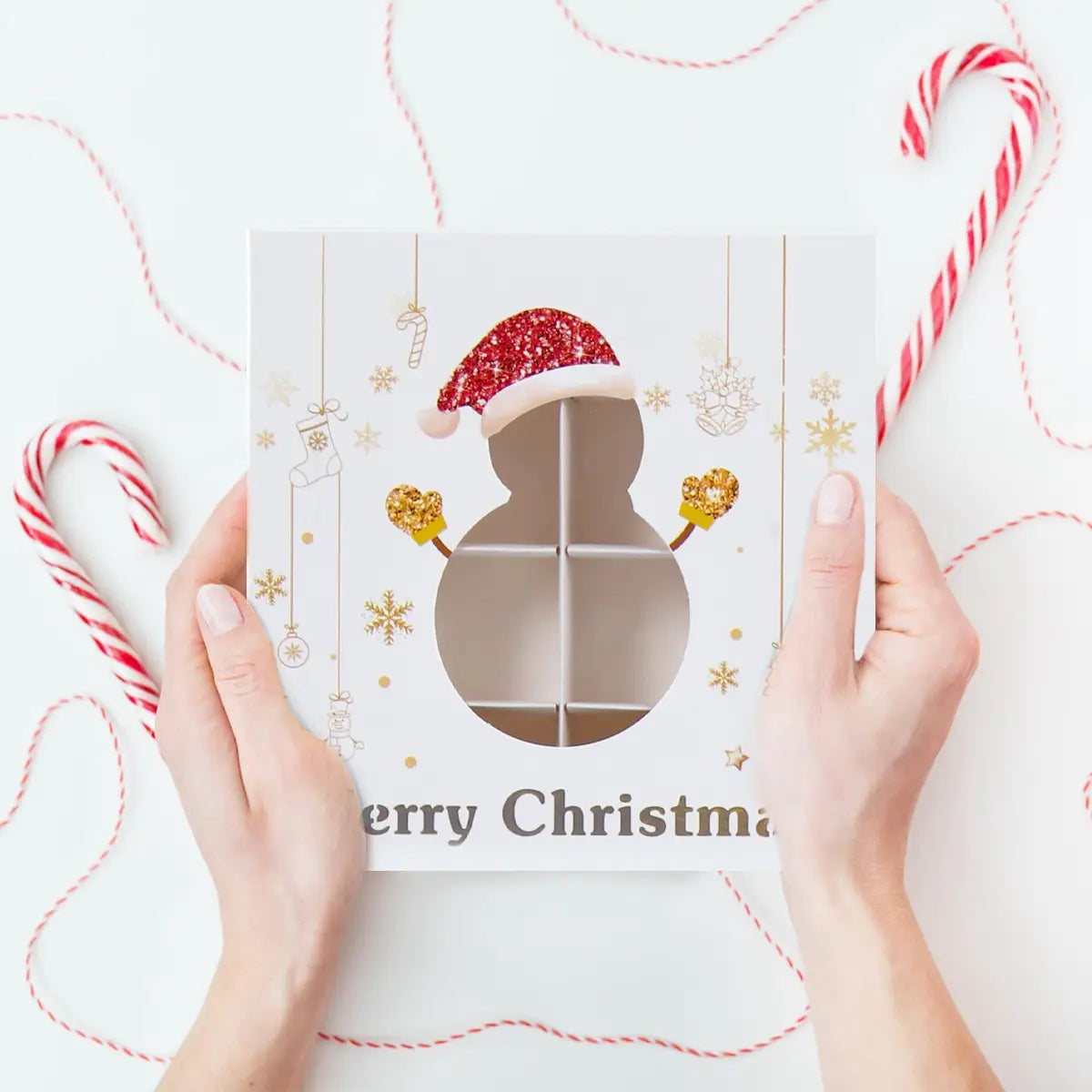 White christmas-themed gift box made of paper with a glittery red santa hat, gold snowflakes, and a clear window divided into six compartments for cookies.