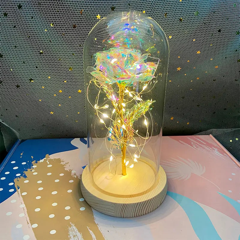 Eine leuchtend irisierende Rose mit goldenem Stiel und zarten Drahtlichtern im Inneren einer klaren Glaskuppel auf einem Holzsockel, im „Eternal Gold Rose Glass Dome LED Light Gift“.