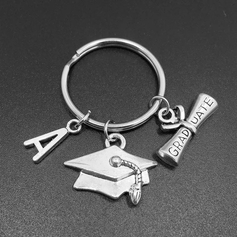 Silbernes Schlüsselanhänger-Geschenk zur Abschlussfeier mit Abzeichen und Quasten-Anhänger, einem gravierten Talisman eines Abschlusszeugnisses sowie einem personalisierten Silber-Anhänger in Form des Buchstabens a.