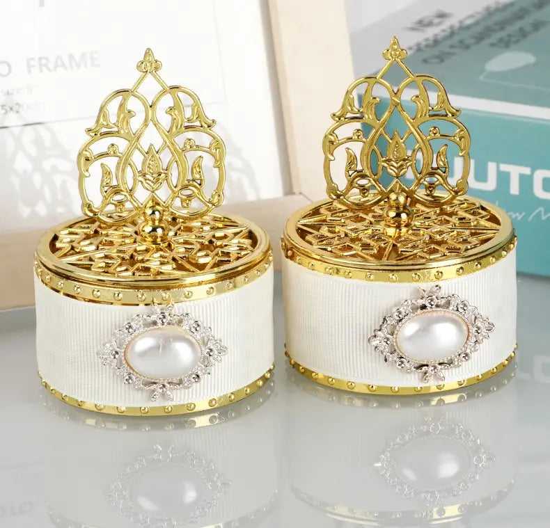 Deux boîtes à bonbons de mariage dorées ornées avec des corps en tissu blanc cannelé et des couvercles bordés d’or, présentant des motifs de filigrane complexes, utilisées comme contenants de perles-bonbon décoratives.