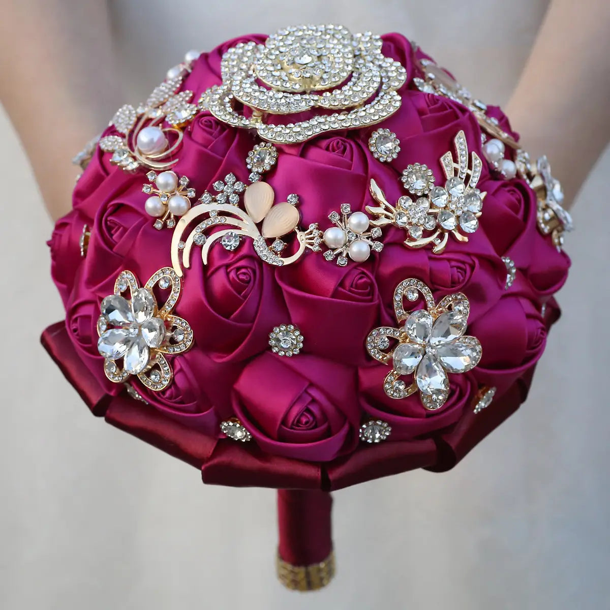 Un luxueux bouquet de mariée en satin fuchsia, rehaussé de strass dorés et de broches en strass étincelantes, d’accents de perles et d’ornements floraux aux tons dorés.