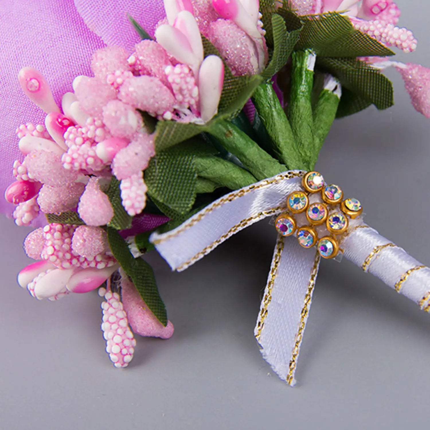 A delicate pink and white floral artificial silk rose boutonniere with sparkling beads green fabric leaves a satin ribbon bow accented by gold trim and clustered rhinestones for blue wedding corsage use.
