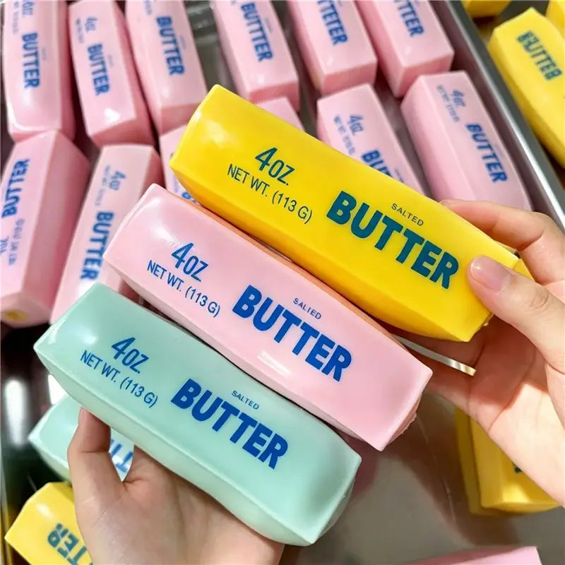 Eine Hand hält einen kräftig gelben 4-oz-gesalzenen Butterriegel als ASMR-Sensory-Squishy-Spielzeug mit kräftiger blauer Beschriftung; daneben sind pinke und mintgrüne Squishy-Spielzeuge im selben Verpackungsstil.