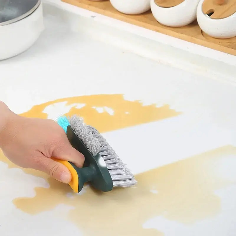 Une brosse de nettoyage en forme de V pour carrelage et rainures avec un manche jaune et vert foncé à double face, présentant des poils gris d’un côté et des poils à pointe bleue de l’autre.