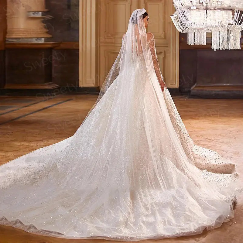 Robe de mariée en style boule perlée, avec des manches longues en tissu blanc fluide, présentant un long voile transparent orné d’ornements étincelants, montrée du côté arrière