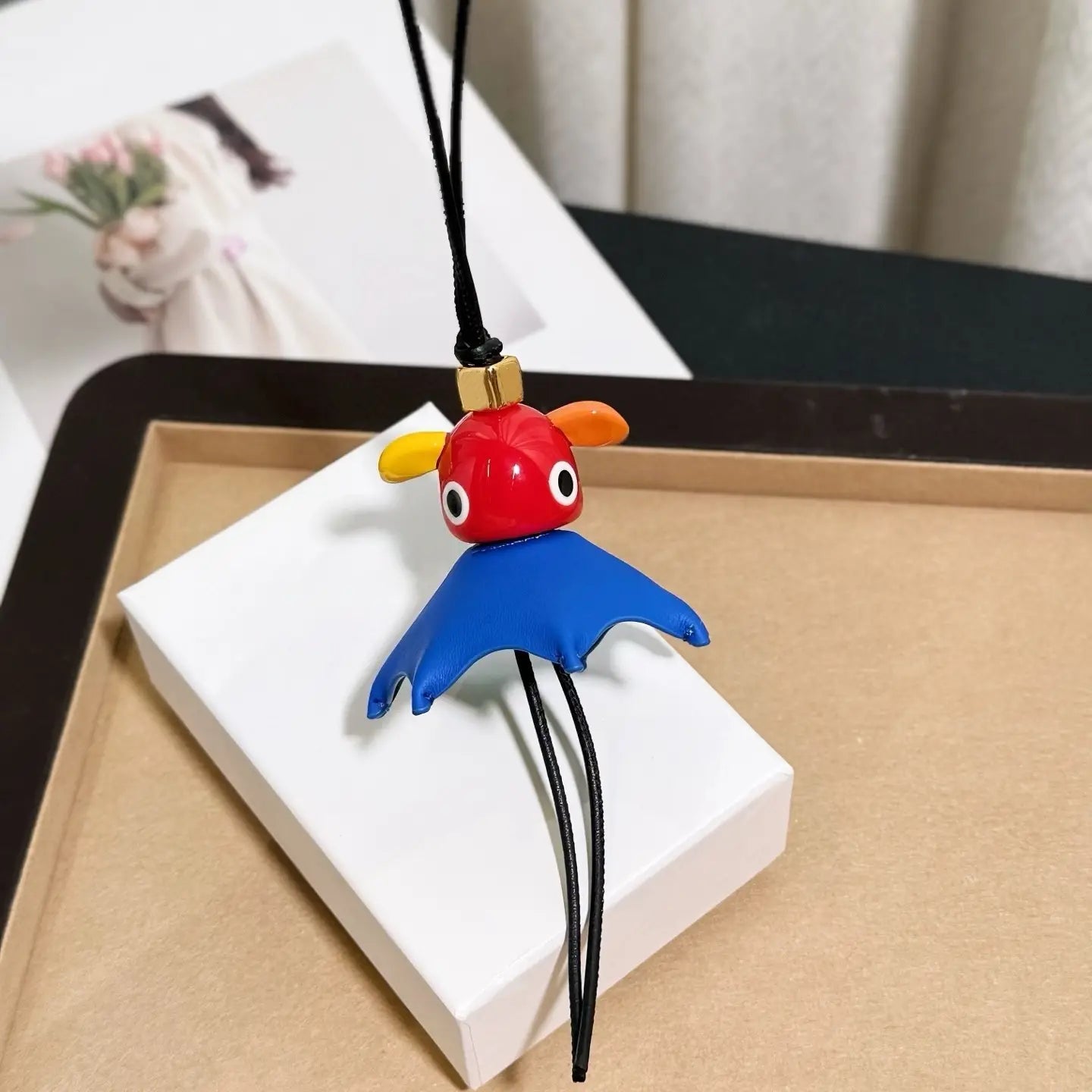 Un llavero colgante para bolso con encanto de animales de cuero rojo y azul vibrante con forma de murciélago caricaturesco, con orejas amarillas, ojos negros y un broche de color dorado diseñado como un llavero colgante de diseñador tierno