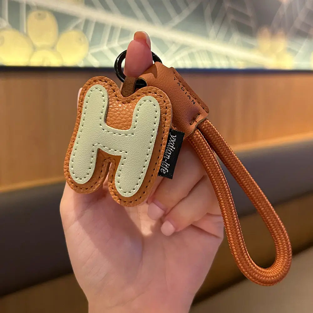 A tan leather initial keychain wristband charm featuring a light beige stitched letter h design with an orange braided strap and black metal ring.