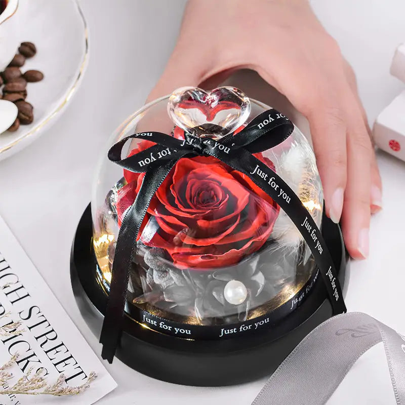 Une rose rouge préservée éclatante enfermée dans un dôme en verre transparent, ornée d’un nœud papillon en ruban noir et d’un cristal en forme de cœur sur une base noire élégante, dans un cadeau en dôme en verre avec rose préservée et éclairage à LED.