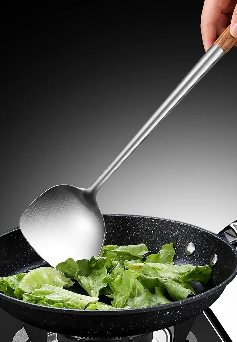 Une élégante spatule à wok en acier inoxydable avec manche en bois, remuant des légumes verts feuillus dans une poêle à frire noire antiadhésive.