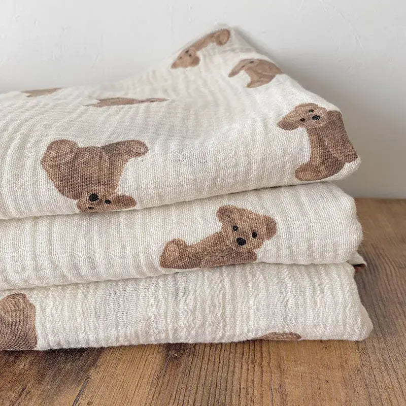 übereinander gelegte Baby-Badehandtücher und Wickeldecken aus beigem Muslin mit einer weichen, strukturierten Webart und bezaubernden Aufdrucken brauner Teddybären