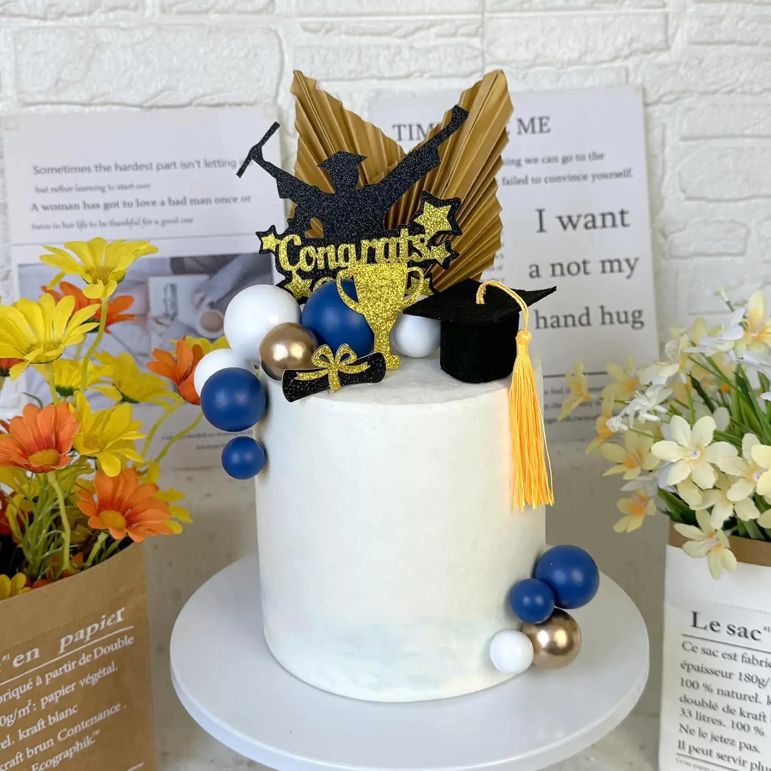 Graduation cake topper set for 2026 senior party featuring a white frosted cake with gold and black glittery topper, blue and white balloons, a miniature black cap with yellow tassel, and a golden trophy decoration.