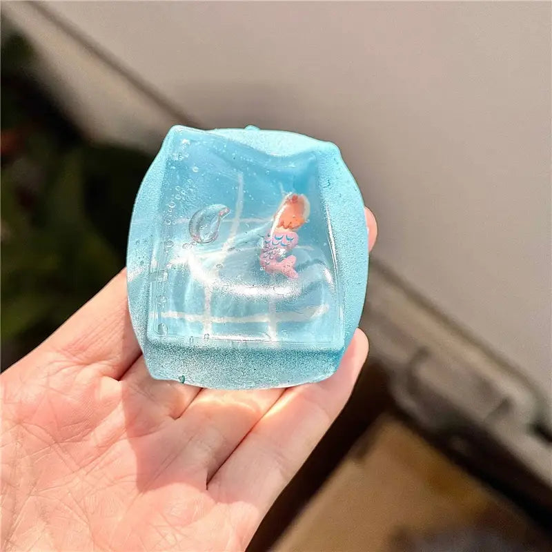 un cube sphérique à faible rebond, visqueux et lent, avec une figurine translucide en résine bleu clair représentant un petit personnage aux cheveux orange et à la tenue rose, enfermé dans un conteneur carré rempli d’un liquide scintillant, vu de face