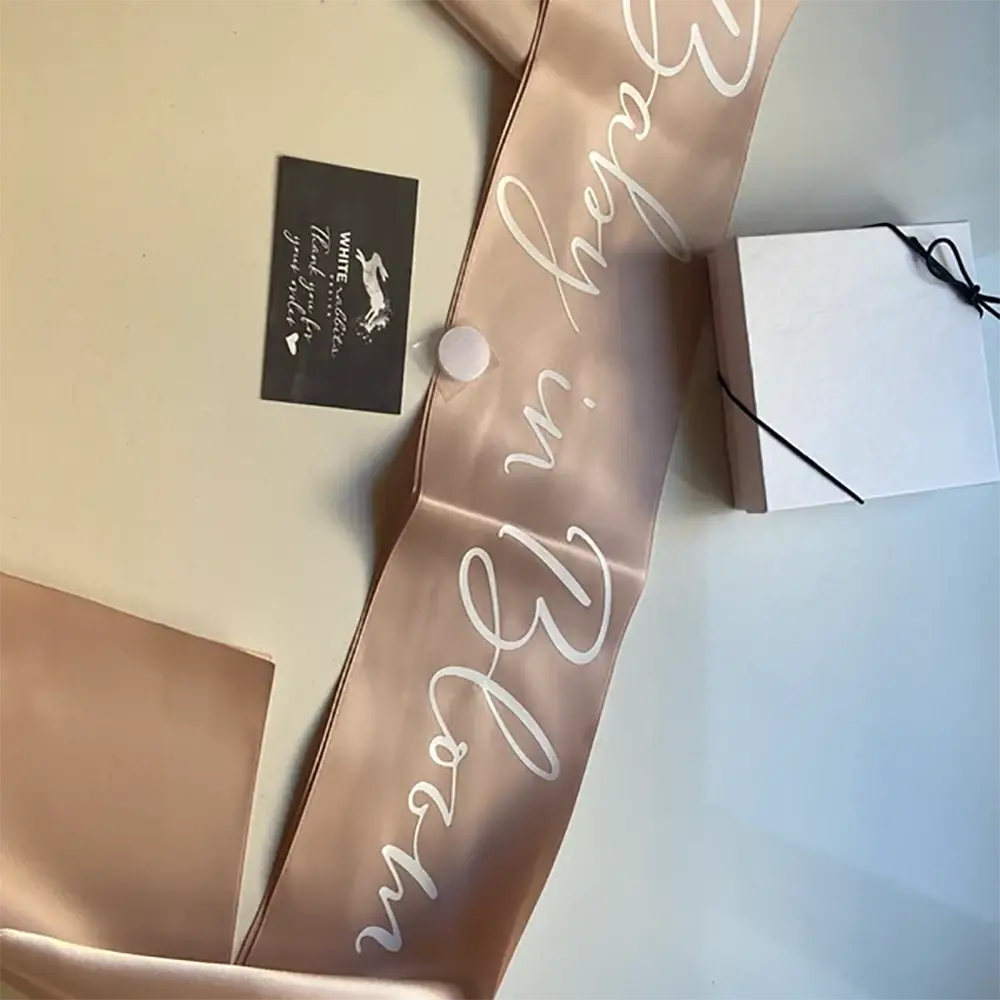 Une écharpe de baby shower en satin rose doré pâle avec une écriture cursive blanche indiquant « baby in bloom », conçue pour la future maman, dans un matériau en satin blush rose pâle.