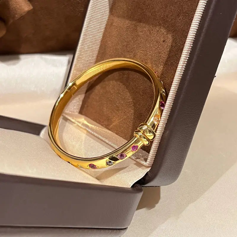 Ein Armreifarmband aus goldfarbenem Multigemsteinschmuck mit einer eleganten, polierten Oberfläche, verziert mit lebhaften Edelsteinen in Pink und Orange.