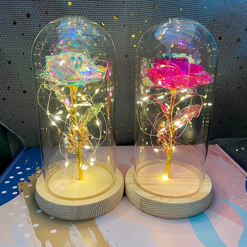 Zwei illuminierte Glasdomenrosen mit einer irisierenden Blütenrose und einer leuchtend pinken Rose, jeweils umschlossen in einer klaren Schutzhülle, mit LED-Licht, das auf einem hölzernen Sockel ruht, im Produkt „Eternal Gold Rose Glass Dome Led Light Gift“