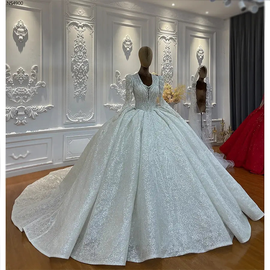 Robe de mariée sur mesure à manches longues en dentelle et jupe en forme de cloche, en sequins argentés scintillants, avec jupe ample présentée sur un mannequin dans une pièce blanche ornée.