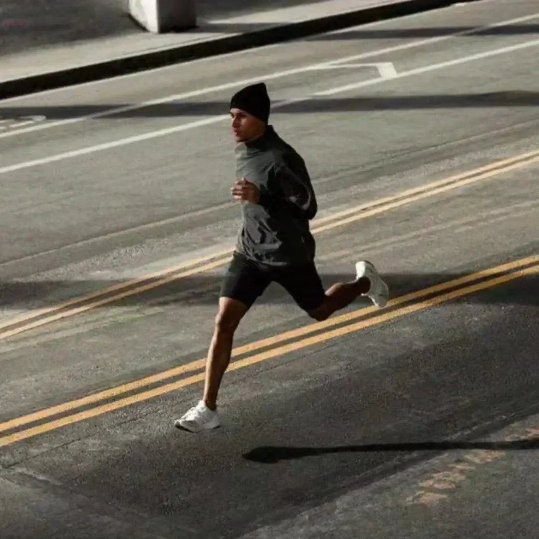 Un hombre en movimiento que lleva pantalones cortos de entrenamiento para anime de gimnasio 2 en 1 para hombres con cintura elástica y trabilla para la toalla, combinado con una gorra tipo beanie negra, parte superior de manga larga gris y zapatillas blancas.