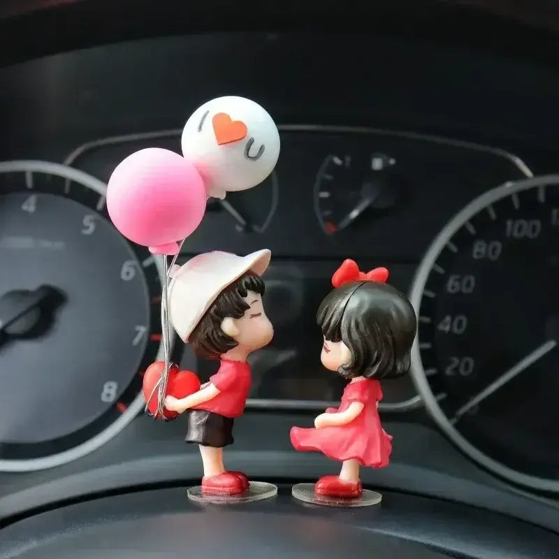 車のダッシュボード上で、ピンクの風船を持つ男の子の赤と白のプラスチック製フィギュアと、ドレスを着た女の子のフィギュアが並んでいる様子をあしらった、かわいいカップルのカー・ダッシュボード用レジンオーナメント。