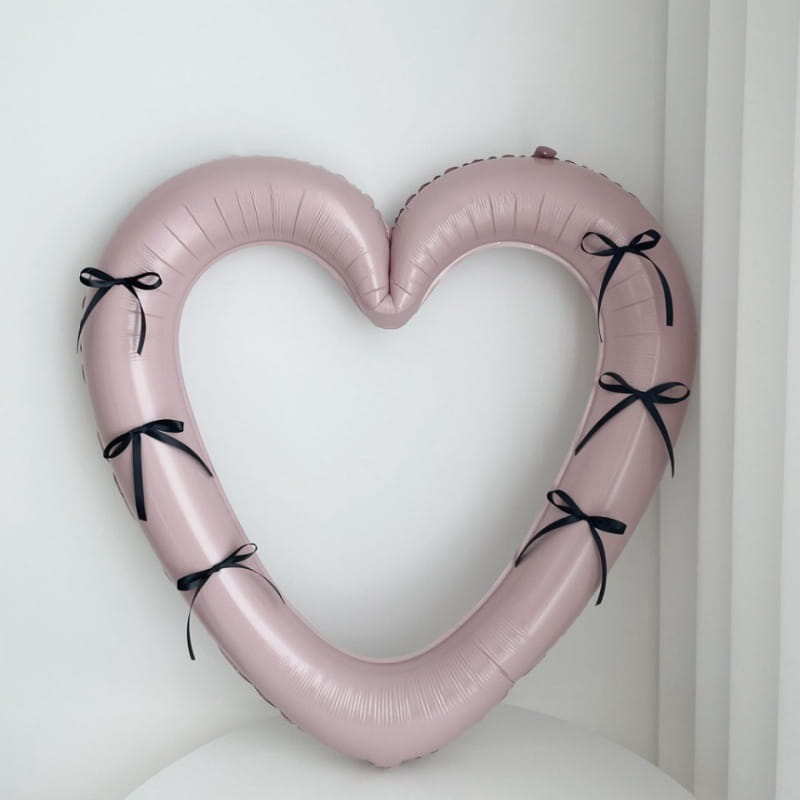Un globo de lámina para marco de foto de corazón de color rosa suave de 40 pulgadas, adornado con moños de cinta negra en los bordes a lo largo de sus curvas.