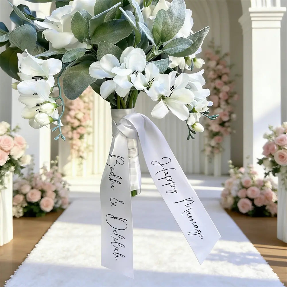 Un ruban de bouquet de mariage en satin blanc personnalisé noué autour d’un bouquet de mariée, présentant un lettrage cursif élégant avec les noms propres bekki et della et la mention « heureuse union ».