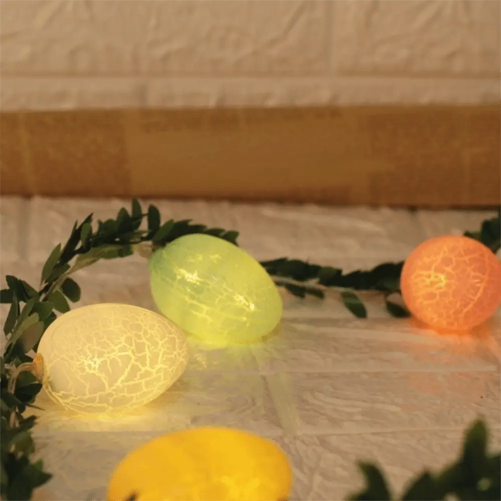 Tres luces LED con forma de huevo brillantes de color amarillo y naranja con diseño de vidrio cuarteado, integradas entre follaje verde en una guirnalda de luz LED tipo huevo de Pascua en ramita de 2 m.